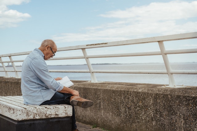An older gentleman is sat outdoors on a bench facing the coast whilst reading a book