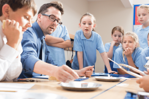 A school teacher is sat down at a school table, surrounded by school children engaging in a practical science lesson and holding equipment